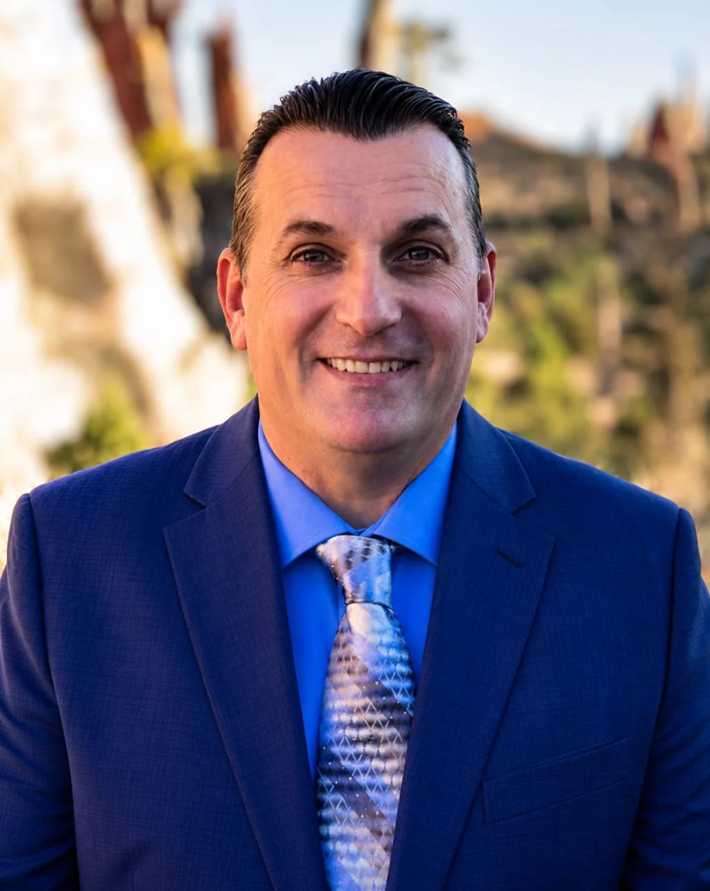 Smiling man in a blue suit and patterned tie, outdoors with a natural rocky background behind him.