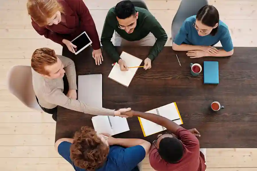 Overhead view of a diverse team around a table shaking hands, signaling collaboration and a deal or agreement.